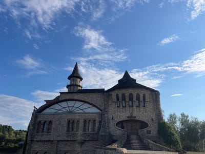 The image shows a unique, castle-like building constructed primarily of stone with conical roofs and turrets. The building has several arched windows and a large arched window with a geometric design on the left side. A statue of a bird of prey is perched on the edge of the roof on the far left. A set of stone stairs leads up to a large wooden door. The sky is blue with scattered white clouds. Trees and foliage are visible to the right and in the background. This building is identified as being located in Colibași, Moldova. No people are visible in the image.