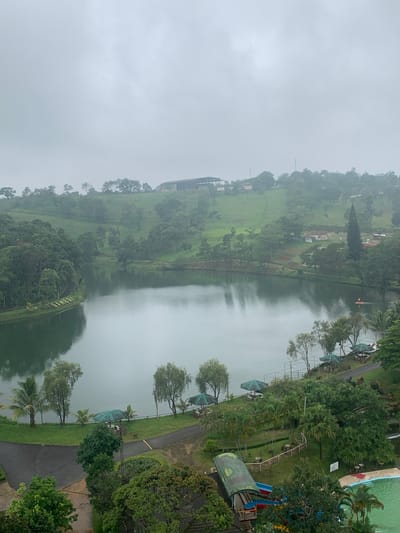 The image shows a scenic view of a lake surrounded by lush green hills and trees. The sky is overcast, indicating a cloudy day. In the foreground, there is a swimming pool with blue and green tiles, and a water slide. There are also several umbrellas providing shade, and some tables and chairs set up outdoors. A paved path winds through the area, and there are various types of trees, including palm trees and other tropical vegetation. On the far side of the lake, there are some buildings nestled into the hillside, and in the distance, more rolling hills covered in greenery. One section of the shoreline has the word "LAMBI" spelled out in what appears to be decorative bushes or plants. A small boat with an orange canopy is visible on the lake. The overall impression is of a tranquil resort or recreational area.