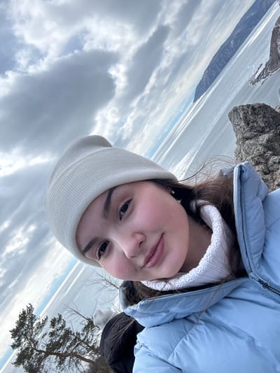 A young female, positioned centrally and slightly angled, occupies the foreground of a selfie-style image taken in Nikola, Russia. She wears a cream-colored knit beanie, a light blue puffer jacket with a visible zipper, and a white high-necked knit garment. Her dark hair is partially visible beneath the beanie, and her cheeks display a flushed appearance. A small, dark earring is visible on her left ear. In the lower-left background, the back of a second individual, wearing a dark jacket and a white beanie, is partially visible. The background features a vast, partially frozen body of water, likely a lake, extending towards a distant, dark landmass or mountain range on the horizon. The immediate foreground and midground include dark, rocky formations on the right and sparse evergreen trees on a rocky incline on the left. The sky is overcast with large grey and white clouds, with intermittent patches of lighter blue sky. The overall scene suggests a cold weather environment.