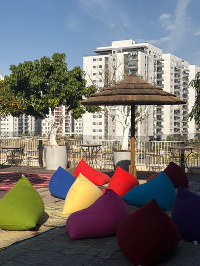 The image depicts an unoccupied outdoor seating area located in Kiryat Ono, Israel. The foreground features a surface covered with woven mats in shades of dark grey, red, and blue-grey. Scattered across these mats are approximately ten brightly colored beanbag chairs in hues of lime green, royal blue, yellow, magenta, red, teal, and burgundy. A large, multi-tiered thatched umbrella with a prominent wooden central pole provides shade in the midground. To the left, a tree with green foliage and orange fruit, its trunk painted white, stands in a large grey planter. Another tree with a white-painted trunk and fewer leaves is visible near the umbrella, also in a grey planter. Beyond the beanbag area, several dark metal round tables and matching chairs are arranged near a white metal railing that delineates the edge of the elevated platform. The background reveals a dense urban environment dominated by multiple tall, modern, light-colored residential buildings featuring numerous balconies. The sky above is clear and blue, indicating a sunny day with distinct shadows cast by the umbrella and beanbags. No individuals are present in the scene.
