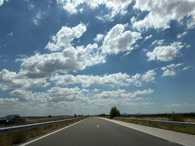 The image depicts an asphalt road with white dashed lines, suggesting it is a highway or a major road. The road stretches into the distance under a bright blue sky filled with fluffy white clouds. On the left side of the road, there is a metal guardrail, and beyond that, a grassy area with some dry vegetation. A dark-colored SUV is parked on the shoulder of the road. The landscape on either side of the road appears to be rural, with fields and some scattered trees visible in the distance. There are no people visible in the image. The scene appears to be a daytime shot under clear weather conditions. The location is identified as being in the vicinity of Krushevo, Bulgaria.