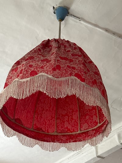 A close-up, upward shot shows a red fabric lampshade with a floral pattern, adorned with white fringe along its lower edge. The lampshade is suspended from the ceiling by a thin metal rod, attached to a blue plastic fixture. The ceiling itself is white and appears textured. The scene is indoors, likely in a residential setting. The location is not discernible from the image itself.