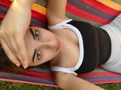 This is a top-down, medium shot of a young woman lying in a hammock, outdoors. She is facing the camera, with her right hand resting on her forehead. Her eyes are open and looking directly at the camera, conveying a sense of calm or relaxation. The woman is wearing a black tank top with white trim and light gray leggings. The hammock she is in has colorful stripes of red, yellow, blue, and green. The background shows green grass, suggesting a natural, outdoor setting. The lighting indicates it is daytime, likely sunny due to the strong shadows and bright highlights. The overall mood is peaceful and serene. No text is visible in the image.