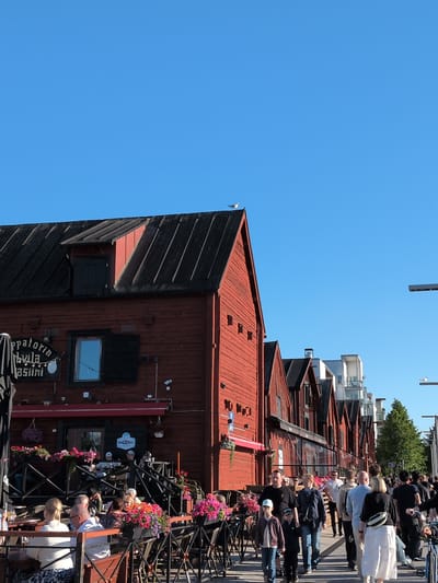 This image depicts a street scene in Oulu, Finland, likely in the marketplace or a similar commercial area, given the presence of shops and outdoor seating. A prominent, weathered red wooden building with a dark metal roof dominates the left side of the frame. The building has several windows, some with dark shutters, and a sign with Finnish text that appears to be advertising a cafe or restaurant. An awning extends over an outdoor seating area in front of the building, where several people are seated at tables.
In the foreground, there are more outdoor seating areas with tables and chairs, adorned with vibrant pink and red flowers in pots. A diverse group of people are walking along the paved sidewalk. Some are walking towards the viewer, while others are walking away. They are dressed in casual summer attire. The background shows a row of similar red wooden buildings interspersed with more modern, multi-story white buildings and some greenery. The sky is a clear, bright blue. A single bird is perched on the roof of the red building on the left. The overall atmosphere suggests a warm, sunny day in a bustling urban environment.