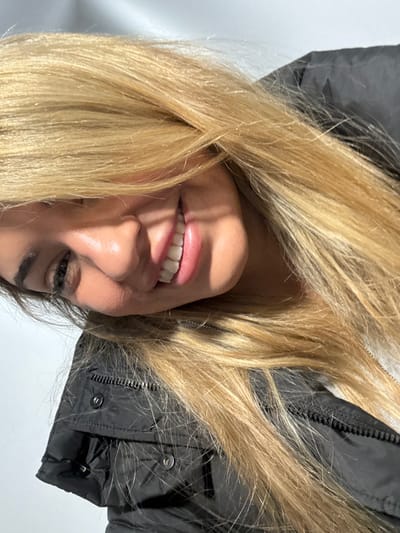 The image depicts a close-up, angled view of a single individual, presumably female, with long blonde hair partially obscuring the face. The person is smiling, revealing upper teeth, with the right eye, nose, and lips clearly visible. The left side of the face is largely covered by hair. The individual is wearing a dark, possibly black, puffer-style jacket with a visible zipper and snap buttons on the collar area. A light-colored surface serves as the background, suggesting an indoor setting or a plain wall outdoors. Harsh, bright light illuminates the subject, casting shadows on the face, particularly under the nose and around the cheekbones. The scene is located in Bad Vilbel, Germany. No other individuals or interactions are observable.