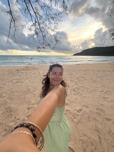 In the city of Preah Sihanouk, Cambodia, a woman with curly brown hair smiles as she looks to her right, holding the camera with her left arm extended forward. She is wearing a light green gingham dress and multiple bracelets on her left wrist, including a gold-colored chain bracelet and a woven brown and black bracelet. She has a pair of sunglasses perched on her head. The woman is standing on a sandy beach with footprints visible in the sand. In the background, gentle waves lap at the shore of a turquoise ocean, and a distant figure is standing in the water. The sky is filled with dramatic, cloudy formations illuminated by a soft, golden sunset behind a dark, tree-covered hill on the horizon. Branches from a tree hang into the frame from the top left.