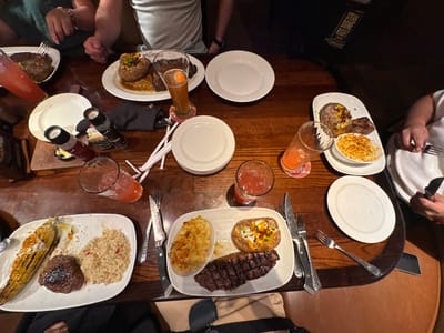 The image captures a dining scene at what appears to be a LongHorn Steakhouse, confirmed by visible text. A dark wooden table is laden with multiple plates of steakhouse fare, suggesting an evening meal due to the warm, dim lighting. Several individuals are partially visible around the table, their hands holding forks and knives as they partake in their meals.
Each diner has a substantial plate, primarily featuring various cuts of grilled steak. Accompaniments include loaded baked potatoes topped with cheese, bacon bits, and chives; creamy mac and cheese in oval ramekins; seasoned rice; and grilled corn on the cob. Beverages include multiple glasses of a pink, possibly fruit-flavored, drink and one tall glass of an amber-colored beer or ale.
White rectangular and round plates, forks, knives with dark handles, and black salt and peppercorn grinders are neatly arranged on the table. Visible text on a bag or menu item clearly states "STEAK DONE RIGHT" and "LongHorn Steakhouse", with smaller labels on the grinders indicating "SALT" and "PEPPERCORNS". The scene depicts a casual American steakhouse in Schaumburg, United States of America, with an atmosphere of people enjoying a hearty meal together.