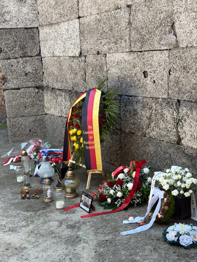 The image depicts a memorial scene, likely at a solemn historical site given the location context of Oświęcim, Poland. The primary subjects are several wreaths of flowers, arranged on a coarse, stony surface. The wreaths are elaborate, with a mix of white, red, and yellow roses, along with greenery and decorative elements like pinecones. Some wreaths are adorned with ribbons, some of which bear visible text. One prominent ribbon, draped across a large wreath, displays text in German: "Der Generalkonsul der Bundesrepublik Deutschland" (The Consul General of the Federal Republic of Germany). Other ribbons have text in what appears to be Slovak, including "SLOVENSKO" and potentially names or dedications.
In addition to the floral tributes, there are several glass memorial lanterns, some containing candles, placed on the ground amongst the wreaths. A small framed photograph, containing multiple portraits, is also visible. The overall setting appears to be outdoors, with rough, hewn stone blocks forming a backdrop or part of the memorial structure. The lighting suggests it is daytime, possibly overcast or with diffused sunlight. The atmosphere conveyed is one of respect and remembrance. The arrangement of the wreaths and lanterns indicates a formal commemoration or tribute.