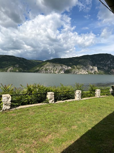 This is a daytime, eye-level, outdoor shot of a wide river flowing between lush, green, and rocky hills under a partly cloudy sky. In the foreground, a well-maintained lawn slopes down towards a low, black metal fence topped with a concrete coping. This fence is supported by four stone pillars, each made of rough-hewn, light-colored stones. Behind the fence, dense green bushes and some taller plants with reddish stems and white blossoms create a natural barrier.

The river itself is calm, with a slightly choppy surface reflecting the sky. The hills on the opposite bank are covered in dense green trees, with exposed rocky outcrops and sheer cliff faces along the water's edge. Several wind turbines are visible atop the hills in the distance. The sky is a mix of dramatic, dark gray clouds and patches of bright blue, with sunlight breaking through in places. The overall impression is one of a serene natural landscape, possibly a scenic viewpoint in Golubac Municipality, Serbia. There are no people visible in the image.