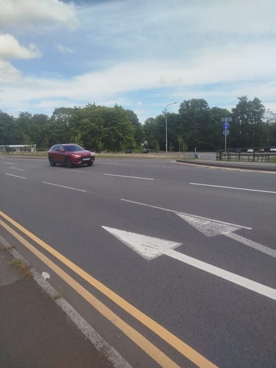 The image shows a street scene in Cwmbran, United Kingdom. In the foreground, there are two white directional arrows painted on the asphalt road, pointing to the left. Double yellow lines are visible along the curb on the left side of the road. A red SUV is driving in the middle lane of the road, facing towards the left. In the background, there are trees, a lamppost, and a bicycle path sign. Several cars are parked on the right side of the road. The sky is blue with some white clouds.