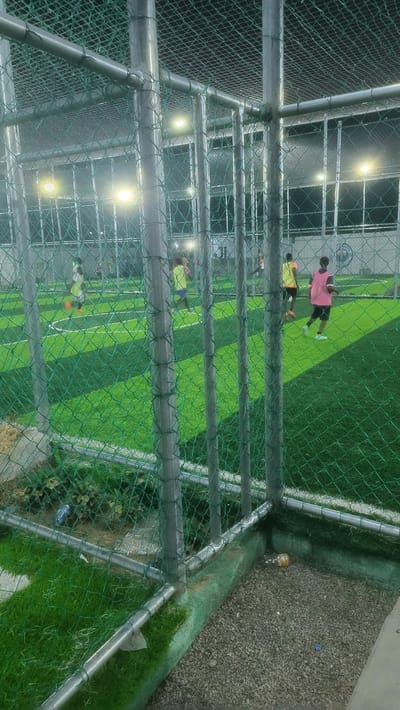 The image displays an artificial turf sports field in Babawa, Nigeria, observed at night through a green chain-link fence supported by grey metal poles and crossbars, which also forms an overhead enclosure. The bright green playing surface features white boundary lines. Approximately 5-7 individuals are on the field, wearing various colored pinnies (yellow, pink) and engaged in active movement, suggesting a game or practice. The field is intensely illuminated by multiple overhead white lights. Outside the fence, a ground area consists of gravel and sections of artificial grass. Debris is present on this outer ground, including a blue plastic bottle in the bottom-left and an orange cylindrical object with a blue bottle cap in the bottom-right. In the background, beyond the field, dimly lit structures with window-like features are visible, and a faint "MANCHESTER CITY" emblem is discernible on a back wall.