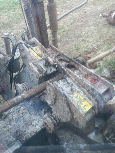The image shows a close-up of a piece of heavy machinery, possibly a drilling rig or similar construction equipment. It is heavily soiled with mud and grime. The machinery is partially outdoors on a patch of grass. There are hydraulic hoses and a cylindrical pipe visible. Specific location in Поедуги, Russia cannot be determined from the provided image. No people are visible.