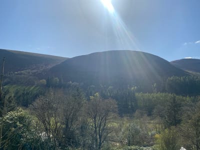 The image depicts a landscape near Llangynog, United Kingdom, on a bright, sunny day. The sky is clear blue with intense sunlight at the top center. Two prominent, rounded hills dominate the midground, with the light illuminating their slopes. The lower slopes of the hills are covered with mixed woodland, including both deciduous and evergreen trees. The foreground is characterized by dense shrubbery and bare trees, with a variety of textures and tones. The image suggests a rural, possibly mountainous environment, with signs of forest and natural vegetation.