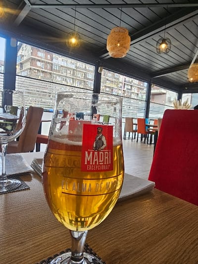 The image depicts an interior scene, likely a restaurant or bar patio, located in Fundeni - Dobroești, Romania. In the immediate foreground, a stemmed glass of "Madri Excepcional" lager beer, approximately three-quarters full with a light foam head and visible condensation, rests on a dark patterned coaster atop a light wooden table. An empty wine glass on a similar coaster is positioned to the left. A portion of a vibrant red upholstered chair is visible on the right.
The midground features additional tables and chairs of varying colors (brown, green, blue, orange) in a blurred arrangement, suggesting an extended seating area. The background, viewed through large transparent window panels, displays an urban residential environment. A multi-story apartment building with balconies and bare trees is distinctly visible.
Overhead, a dark, possibly slatted or paneled ceiling is equipped with multiple pendant light fixtures, including two black wire-cage designs and two natural-toned woven rattan-style designs. No individuals are present in the frame.