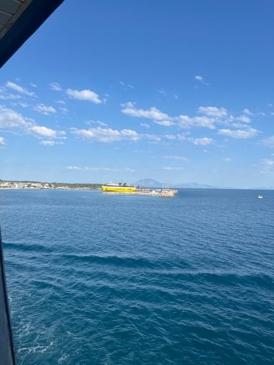 A bright yellow ferry with "Levante Ferries 11880" written on its side is docked near a breakwater in the Municipal Unit of Kastro - Kyllini, Greece. The ferry is positioned to the right of a coastal town with white buildings and scattered trees. In the distance, hills rise from the horizon. The sea is a deep blue, with small waves rippling across its surface. The sky is a clear blue, dotted with fluffy white clouds. A small white boat with a green trim is visible on the water to the far right. The image is framed by a dark, angled structure on the left side.
