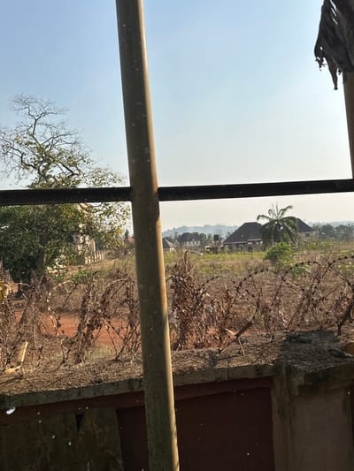 This image depicts an outdoor scene in Agbogugu, Nigeria, characterized by a mix of undeveloped land and residential structures under a hazy daytime sky.
In the immediate foreground, a concrete perimeter wall, approximately 0.5 meters high, is visible. It is topped with multiple strands of coiled concertina razor wire, showing signs of rust and weathering. A vertical, cylindrical, light-colored metal pole, approximately 10-15 cm in diameter, extends from the bottom of the frame to the top, positioned slightly right of center. A horizontal, dark, rusty metal bar runs across the mid-lower frame, connected to this vertical pole. A dark, possibly metal, surface is visible at the bottom edge, behind the concrete wall.
Beyond the immediate foreground, the terrain consists of dry, reddish-brown earth with sparse, desiccated vegetation. Further back, denser green foliage is present. In the mid-ground to background, several residential buildings with dark-tiled, hip roofs are scattered across the landscape. One prominent large house with a dark roof stands slightly right of center. To its left, another large house is visible. A tall, largely leafless tree with intricate branching patterns dominates the left side of the frame, with some green foliage at its base. A distinct palm tree stands to the right of the central large house.
In the distant background, a line of hazy hills or mountains is faintly visible against a light blue, hazy sky. No people, specific actions, or interactions are discernible. The scene is illuminated by strong natural light, casting no distinct shadows due to the direct angle or atmospheric haze.