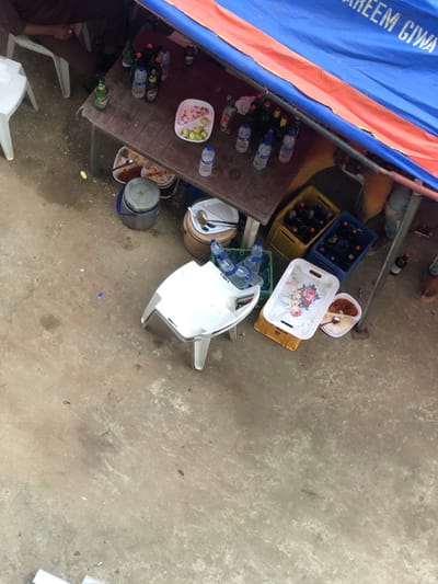 This image, taken in Aiyetoto-Asogun, Nigeria, presents an overhead view of an outdoor, informal gathering area under a temporary blue and orange tarpaulin labeled "AREEM GIWA". A dark brown rectangular table is central, laden with multiple plastic bottles of water, various dark glass beverage bottles (some with green labels, like "Tiger"), smaller clear and dark bottles, and a white rectangular plate containing cut green and red fruit segments.

Below the table, on a light grey concrete or packed earth surface, are several items: a grey lidded plastic bucket, a brown plastic bucket holding white plates and serving utensils, and multiple food containers. These include two white rectangular trays with floral patterns, one presenting a bowl of red stew-like food with a spoon, and other bowls containing food. Four plastic crates (two yellow, two blue) filled with dark glass bottles are positioned on the ground. Three white plastic chairs are visible; one is partially occupied by an individual wearing a dark maroon shirt and trousers, another holds plastic water bottles, and a third is empty. The lower body of a second individual, wearing light brown trousers and brown shoes, is visible on the right, near a single dark bottle on the ground.