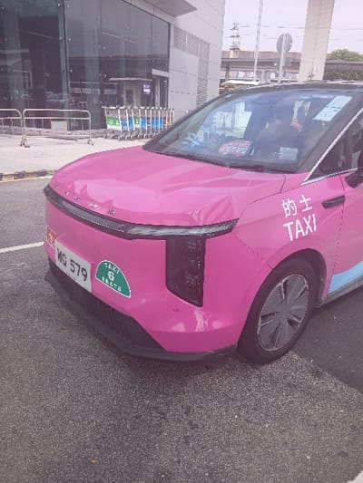 The image displays a bright pink taxi, identified as a 6-seat vehicle with the license plate MG 579, parked on an asphalt surface in Tung Chung, China. The vehicle, possibly a BYD electric model, features "的士 TAXI" in white lettering on its front passenger door and a green and white "TAXI 6 SEATS" sticker on its front bumper. A faint blue stripe runs along the lower side of the vehicle. A person is seated in the driver's position, partially visible through the windshield. The vehicle's front hood exhibits minor surface deformation above the license plate, and slight creasing is visible on the driver's side front fender. In the background to the left, a modern building with large glass windows reflects elements of the surroundings, including a line of silver and blue airport luggage trolleys and metal crowd barriers. An elevated concrete structure, likely an overpass or part of a railway system, is visible further in the background to the right. The scene is illuminated by daylight under what appears to be an overcast sky.