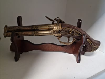 The image shows a decorative, double-barreled flintlock pistol resting on a wooden stand. The pistol is ornately decorated with brass inlays and has a wooden grip. The stand is made of dark wood and has a simple, functional design. The background is a plain white surface, likely a shelf or table, against a neutral-colored wall. The lighting is soft, casting gentle shadows. There are no people visible in the image. The setting does not provide any specific indicators of its location within Pogranichny, Russia; it appears to be an indoor display.