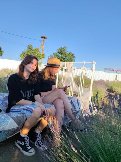 Two young women are seated on a patchwork quilt spread on the ground, positioned in front of a white ornate bed frame. The woman in the foreground, on the left, has light brown hair and is wearing a black t-shirt with white text and blue and red striped shorts. She is looking towards the camera with a slight smile. The woman behind her, on the right, is wearing a straw hat and a black t-shirt. She is looking down at her phone.
The scene appears to be a lavender field, with rows of blooming lavender plants visible in the background. A white picket fence runs along the perimeter of the field, and trees are scattered beyond it. In the distance, a stork's nest is visible atop a wooden pole. The sky is a clear, bright blue, suggesting it is daytime. The lighting indicates that the sun is low in the sky, casting long shadows and creating a warm glow, suggesting it is either early morning or late afternoon.
The overall mood is relaxed and peaceful, with the women enjoying their time in the scenic setting. The visible text on the t-shirt of the woman in the foreground reads "Type". The location is identified as Nechaiane, Ukraine.