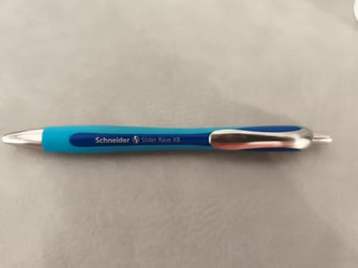 A blue and silver retractable pen, identified by the text "Schneider Slider Rave XB" printed in white and blue on the barrel, lies horizontally on a textured gray surface. The pen is positioned in the center of the frame, with its tip pointing to the left. The surface appears to be a fabric or faux leather material, possibly a desk mat or the surface of a piece of furniture. The lighting is neutral, casting soft shadows that highlight the pen's metallic clip and the texture of the surface. No people or other objects are visible in the image. The location in the city of Essenbach, Germany, is not discernible from the provided image.