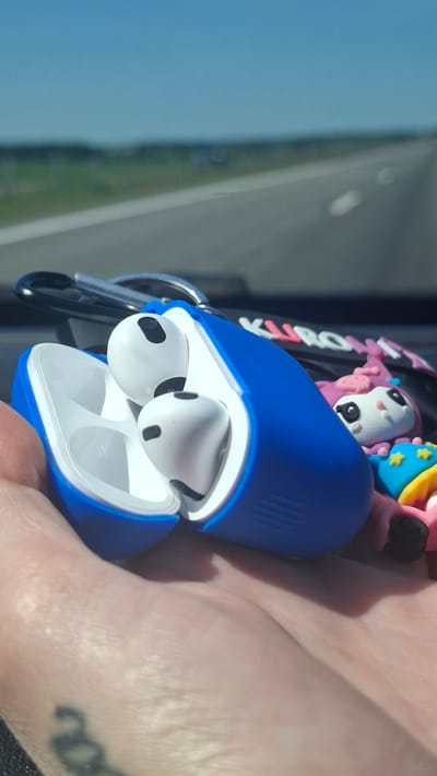An over-the-shoulder shot shows a hand holding a blue case with white earbuds inside. The case is open, revealing the earbuds and their charging slots. A keychain with cartoon characters and the word "KUROMI" is attached to the case. In the background, a road is visible with a green field and blue sky. The image appears to have been taken in a car, possibly on a highway. The scene is lit by natural sunlight. No specific location in Станькаўскі сельскі Савет, Belarus is identifiable.