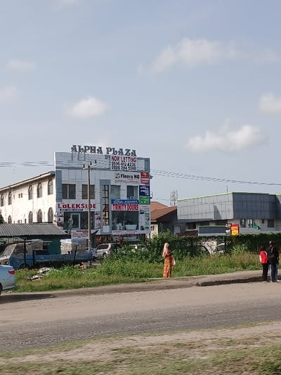 The image shows a street scene in the city of Road, Nigeria, located at the coordinates 6.439° N, 3.52° E. The scene features a multi-story building with a sign that reads "ALPHA PLAZA" and "NOW LETTING," alongside various smaller businesses, including "LOLESKIDE" and "EXTRA POWER TRINITY DOORS." There are two women standing on the street in front of the building. The woman on the left is wearing a hijab, while the woman on the right is wearing a red top. A blue truck is parked in front of the building. There is a large amount of vegetation on either side of the street. The sky is a pale blue with some white clouds.
