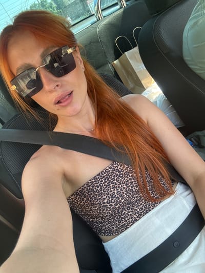 The image shows a woman with long, auburn hair sitting in a car. She is wearing a leopard-print top and sunglasses. A seatbelt is visible across her body. The interior of the car is dark, with black seats. A brown paper bag with handles is visible on the passenger seat. Outside the car, a blurred view of buildings and trees can be seen through the window, suggesting an urban or suburban setting during the daytime. The woman's mouth is slightly open, and her expression is neutral. The image is taken from a high-angle perspective, looking down at the woman. The lighting suggests it is daytime. The text "www.terza.io.com" and "@terzainiofficial" is visible on the paper bag.