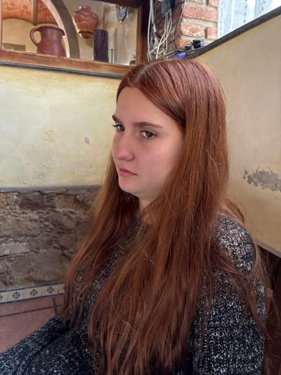 A close-up, slightly angled view shows a woman with long, reddish-brown hair, wearing a speckled gray and white sweater. Her gaze is directed downwards and slightly to the left. The background reveals details of a building interior. Behind the woman and to the right, there's a wall with a rough, stone-like texture. To the left, a framed display contains pottery items. The architectural style of the surroundings suggests the photo was taken in Tepeji de Ocampo, Mexico. Additional visible elements are electrical wires and some equipment near the upper left corner, above the framed display. The lighting appears to be natural.