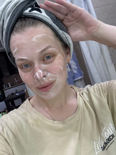 The image shows a young woman with a facial mask on her face. She is wearing a light brown long-sleeved shirt with "LIVE TOUR MCMXCII UNITED STATES" printed on it in white text. A thin silver chain necklace is visible around her neck. Her hair is dark and pulled back, covered by a grey and black striped towel or headwrap. A delicate silver ring with a small stone is on her left ring finger, and a silver bracelet with circular charms is on her right wrist. Her eyes are blue and wide open, looking directly at the camera. The scene appears to be indoors, possibly a bathroom or bedroom, with a tiled floor and what looks like a shower curtain in the background. There are some shelves with bottles visible in the blurry background. The lighting suggests daytime, and the overall mood seems relaxed and informal. The woman's expression is neutral.