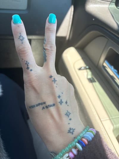 A person's hand, adorned with tattoos and turquoise nail polish, is making a peace sign inside a car. The tattoos are of small stars and the phrase "Wish Upon A Star". The person is wearing colorful beaded bracelets. The interior of the car is beige. The location cannot be determined from the image.