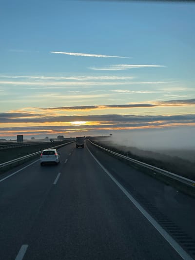 A slightly angled view of a multi-lane highway at sunrise. A white car and a truck are visible, traveling on the road. The sky is a gradient of blue to orange, streaked with long, thin clouds and contrails. Patches of fog are visible along the edge of the highway. The highway's shoulder has guardrails.