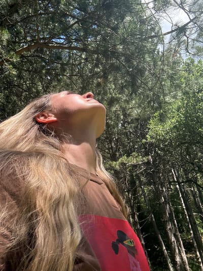 A young woman with long blonde hair is shown from a low angle, looking up towards the sky amidst a dense forest. Her face is illuminated by sunlight, highlighting her features as she tilts her head back. She is wearing a brown long-sleeved shirt with a red graphic print on the left sleeve. The background consists of tall, slender trees with dark bark and green foliage, creating a natural, outdoor setting. The overall impression is one of peacefulness and connection with nature. This scene is located in the village of Žlijebi, Montenegro.