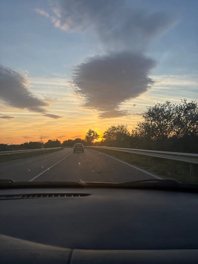 The image depicts a view from inside a car driving on a highway. The scene is oriented vertically. The sky is visible through the windshield, with a mix of blue sky and clouds illuminated by the setting sun, creating an orange and yellow glow. The sun is positioned at the end of the road, obscured by trees. There is a car ahead on the road. The dashboard and side of the car interior are visible on the right side of the image. The location cannot be determined based on the information in this image. No people or interactions are visible.