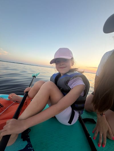 A young girl, wearing a pink baseball cap and a life vest, sits on a paddleboard in the calm waters of a lake. The sun is low in the sky, casting a warm glow on the scene. The girl is holding a paddle, and her bare feet are visible. Another person's arm and hand, with bright pink nail polish, are visible on the right side of the image. The paddleboard is turquoise and orange. The backdrop is a clear sky with a hint of pink and orange from the sunset, and a distant treeline. The scene appears to be in Salaspils, Latvia, during a peaceful sunset on the water.