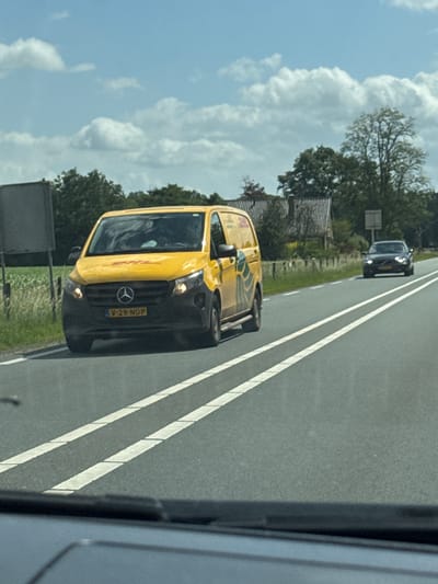 The image captures a view from inside a vehicle traveling on a road in Wierden, Netherlands. A yellow DHL delivery van is prominently positioned on the road, in motion. A black car is visible further ahead on the right lane. The surroundings include roadside vegetation, trees, and a building with a tiled roof in the background. The sky is partly cloudy. The van's license plate reads "V-29-NDP".