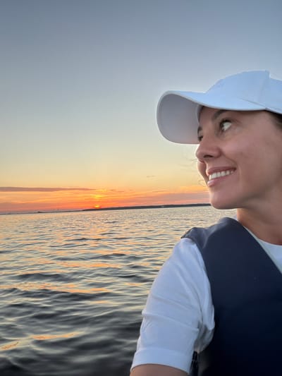The image shows a woman wearing a white baseball cap and a dark vest over a white t-shirt, smiling and looking to the right. She is on a body of water, likely on a boat, as indicated by the rippling water surface. The background features a sunset with orange and yellow hues illuminating the sky and reflecting on the water. The horizon shows a faint silhouette of a distant landmass with some structures. The scene appears to be a peaceful moment captured during the golden hour. The location is identified as Salaspils, Latvia, suggesting the water body could be the Daugava River or one of the lakes in the vicinity.