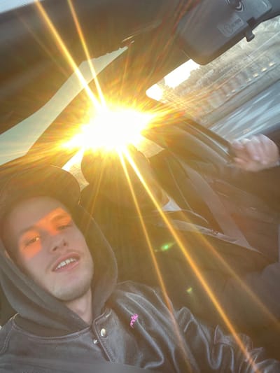 The scene depicts the interior of a vehicle, implied to be located in Caluire-et-Cuire, France. A male individual is prominently featured in the foreground, facing the camera, suggesting a selfie or direct photograph. He wears a dark hooded jacket, potentially leather, with metallic buttons and a pink embroidered insignia on the left chest, and a dark baseball cap beneath the hood. He has short facial hair on his chin and upper lip, and his mouth is slightly open. Intense sunlight streams through the front windshield, creating significant glare and lens flares, directly illuminating the left side of the foreground individual's face. In the background, partially obscured by the solar glare, the right arm and hand of a second individual are visible, gripping an interior element, possibly a door handle or vehicle frame. The strong light indicates bright outdoor conditions, likely sunrise or sunset.