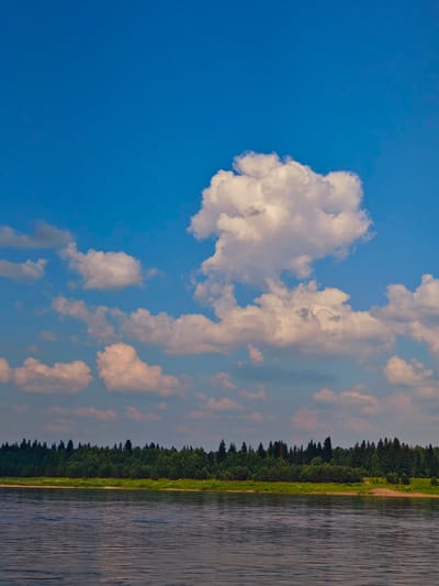 The image depicts a serene natural landscape with a wide, calm river in the foreground. The water surface exhibits subtle ripples and reflections, suggesting a gentle breeze. Along the opposite bank of the river, a dense, verdant forest stretches horizontally across the frame, comprising a variety of evergreen and deciduous trees. The forest line is thick and unbroken, creating a natural barrier. Above the forest, a clear blue sky dominates the upper portion of the image. Several fluffy white cumulus clouds are scattered across the sky, adding texture and depth. The lighting indicates it is daytime, likely late morning or early afternoon, with the sun illuminating the clouds from the side, casting soft shadows. There are no visible human figures, structures, or any signs of urban development. The scene is one of unspoiled nature, characteristic of the rural or wilderness areas found in regions like Ust'-Kem', Russia, known for its vast forests and rivers.