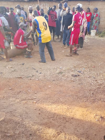 A group of people gather outdoors. One person, wearing a red jersey, sits on a wooden block with chains around their ankles, holding hands with a person in a yellow jersey. Other individuals stand around, some holding bowls or other items. The background shows a wall with some graffiti or markings. The ground is dirt.