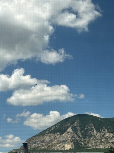 This media file captures a scenic view of a steep, vegetated mountainside meeting a clear blue sky dotted with fluffy white clouds. The mountain on the right is rugged and appears to be covered in trees and foliage, with patches of exposed rock. A narrow, dark-colored structure, possibly a fence or guardrail, runs along the edge of the mountain, suggesting a road or viewpoint. In the sky, there are several cumulus clouds, some appearing soft and wispy, while others are more dense and voluminous. The lighting indicates it is daytime, likely mid-morning to afternoon, with the sun casting light on the mountain and illuminating the clouds. There are at least two dark, bird-like figures visible in the sky, suggesting the presence of wildlife, possibly birds of prey due to the mountainous environment. The overall impression is one of natural beauty and vastness, with the imposing mountain juxtaposed against the open sky. The image is oriented vertically, with the mountain occupying the right side and the sky the left. There is no visible text within the frame.