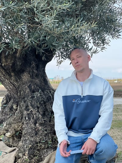 A man sits on a large olive tree in an outdoor setting in Зеленогорское сельское поселение, Ukraine. He wears a two-tone white and navy blue jacket with white sleeves and a white collar, blue jeans, and a white t-shirt underneath the jacket. He holds a small white package in his hands. The olive tree is gnarled and thick, with a dense canopy of green leaves. The background shows glimpses of a rural or semi-rural landscape.