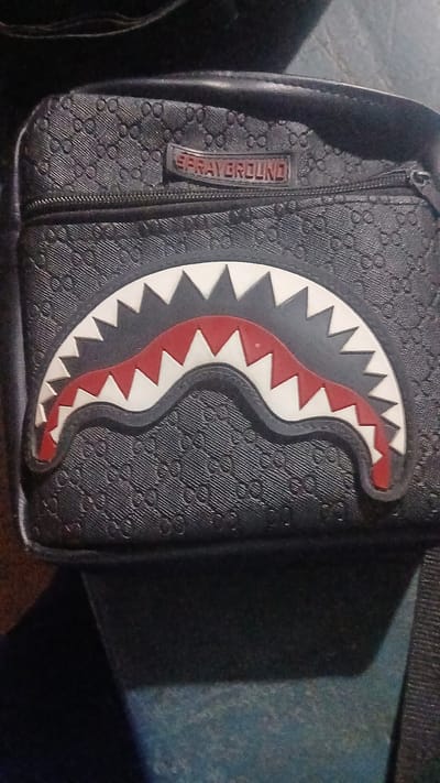 The image shows a black Sprayground shoulder bag with a shark mouth design on the front. The shark mouth is white with red accents and black outlines. The bag has a patterned texture, possibly a repeating logo. A zipper is visible on the side of the bag next to the "SPRAYGROUND" logo. The background is blurry and dark, and no specific details suggest a location. The location in Putucual, Venezuela is not discernible in the image.