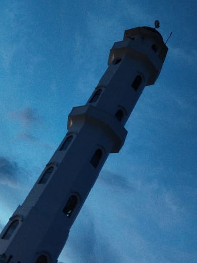 A light-colored minaret, likely part of a mosque, stands tall against a clear blue sky. The minaret is octagonal in shape and features multiple arched windows on each level. At the very top, a crescent moon symbol is visible. The minaret is angled to the right, creating a dynamic composition against the expansive sky. The lighting suggests it is either dusk or dawn, as the sky has a deep blue hue. The overall impression is one of serene verticality and religious symbolism.