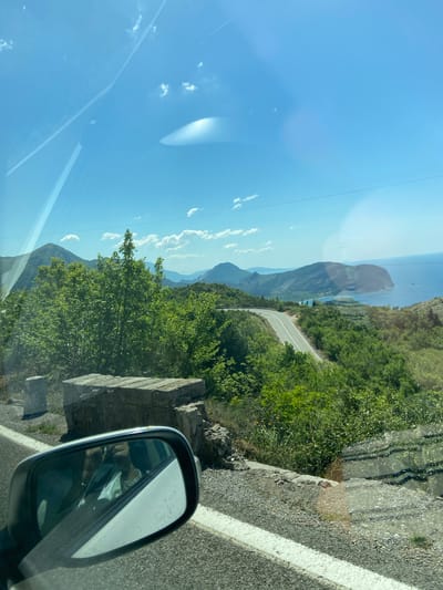The image captures a view from inside a vehicle, likely a car, traveling on a mountain road. The location is suggested by the query to be in the city of Nerin, Montenegro. The road winds through a mountainous landscape with lush green vegetation. In the distance, there's a view of the sea. The sky is clear and blue with a few scattered clouds. The car's side mirror is visible in the foreground, reflecting the interior of the car. The road has a white line marking the edge. The image has a slight upward angle and appears to have been taken through a window, as evidenced by the reflections and artifacts in the blue sky.
