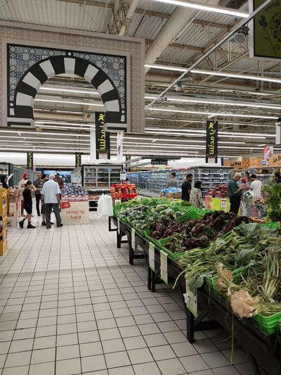 The image depicts the interior of a supermarket in Cebalat, Tunisia. The scene is dominated by aisles filled with produce and shoppers. In the foreground, a wide variety of fresh vegetables are displayed in green crates on dark wooden stands. These vegetables include beets, leafy greens, and root vegetables with their tops still attached. Shoppers, dressed in casual attire, are seen browsing the produce section and interacting with each other.

Further back in the supermarket, aisles are lined with refrigerated display cases stocked with food items. Several banners with Arabic script hang from the ceiling, indicating different sections or promotions. An ornate archway with a black and white striped pattern and intricate blue and white tiling is visible on the left side of the image, suggesting a themed or decorative element within the store. The lighting appears to be fluorescent, typical of a commercial environment. The floor is tiled with a light-colored grid pattern. The overall impression is of a bustling marketplace within a retail setting.