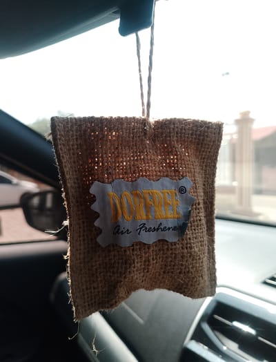 A burlap air freshener hangs from the rearview mirror of a car, the interior of which is visible. The air freshener is light brown and rectangular, with frayed edges. A light blue tag with a scalloped edge is attached to the front of the air freshener. The tag has yellow text that reads "DORFREE" and black text below it that reads "Air Freshener." The background is a bright, out-of-focus view of what appears to be a sunny outdoor scene. The car's interior is dark, with a glimpse of a black dashboard and a light-colored seat. The scene is indoors, within a vehicle. No people are visible. The image is taken in Meunasahuket, Indonesia.