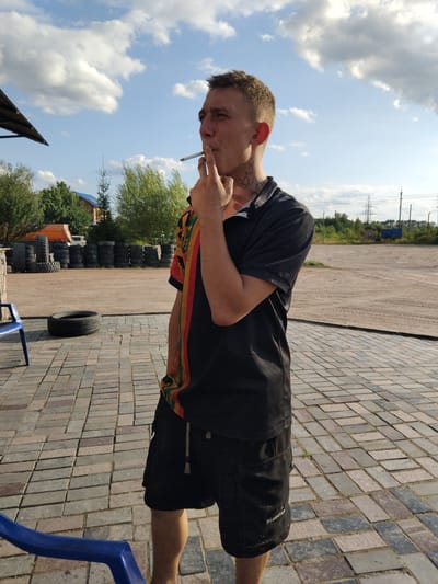 In Novoselytsia, Ukraine, a young man with short, light brown hair stands outdoors on a paved area with brick-like stones. He is wearing a black polo shirt with colorful accents on the sleeve and black cargo shorts. The man is smoking a cigarette, holding it to his mouth with his right hand. He has tattoos on his neck. In the background, there is a stack of tires, some trees, a building, and power lines against a blue sky with scattered clouds. A blue plastic chair is partially visible in the lower left corner of the image.
