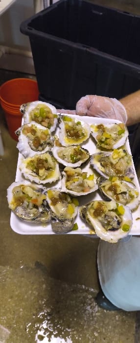 A right hand, encased in a translucent disposable glove, holds a white styrofoam tray containing twelve oysters on the half shell. Each oyster is topped with a mixture of finely diced green and yellow vegetables, possibly onions or peppers, along with a brownish-gold sauce or dressing, and what appears to be a clear liquid or oil. In the upper background, a large, black, rectangular plastic bin is visible. To the left, a portion of an orange bucket is present. The lower part of the image reveals a wet, light-colored concrete floor with visible water and scattered particulate matter. The scene is located in Cutler Bay, United States of America.