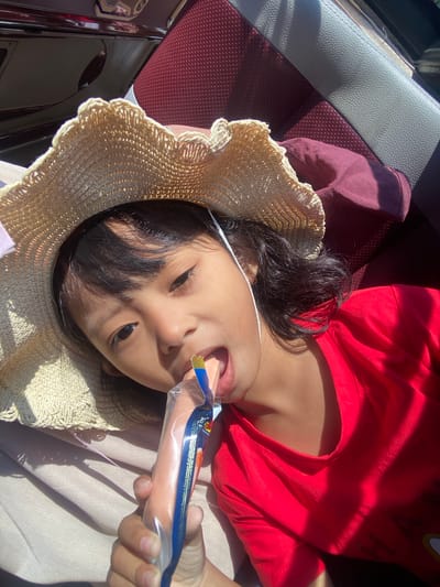 A young child, likely of Southeast Asian descent, is observed inside a vehicle. The child is wearing a woven straw hat with a light-colored ribbon and a red short-sleeved t-shirt. They are actively consuming a processed food item, specifically a pinkish, elongated snack encased in clear plastic packaging with blue and yellow accents, which they hold in their right hand and bite into. The text on the packaging is partially visible and appears to be in Indonesian. Strong direct sunlight casts defined shadows across the scene, illuminating the child's face and clothing, and highlighting textures within the vehicle's interior. The background features elements of a vehicle's interior, including a textured maroon seat and a lighter gray seat component. The child's head rests on a light-colored fabric. The scene is located in Sumber Suko, Indonesia.